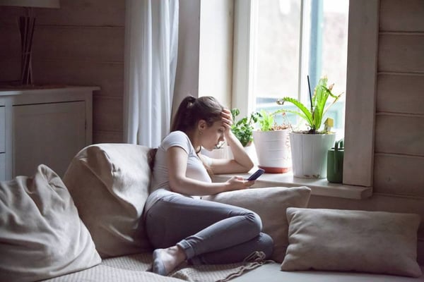 Ung tjej sitter i soffa med handen mot pannan och tittar på mobilen